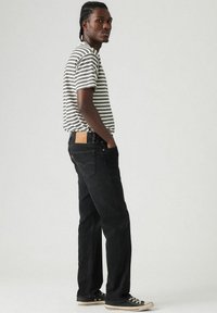Zwarte jeans met een leren patch op de achterkant, gecombineerd met een gestreept zwart-wit T-shirt. Model staat zijwaarts en draagt sneakers.