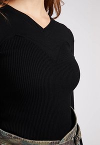 Pull noir côtelé avec un décolleté en V, arborant des détails en chevron texturé sur la poitrine. Associé à une ceinture camouflage à motifs.