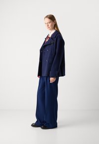 Chaqueta doble botonadura de lana en color marino con un bolsillo, que presenta un lazo decorativo y una camisa crema de contraste, combinada con pantalones anchos en tono marino.