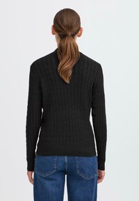 Schwarzer Strickpullover mit strukturiertem Zopfmuster. Figurumspielendes Design, lange Ärmel, gerippter Saum. Kombiniert mit blauen, hoch taillierten Jeans.