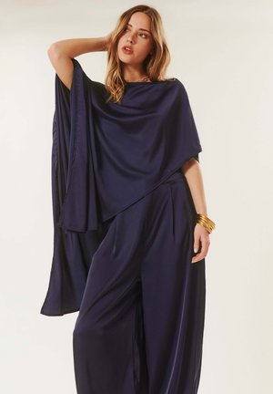 Femme portant un pantalon large bleu marine foncé et un haut asymétrique style poncho assorti, accessoirisée avec des bracelets dorés à un bras.