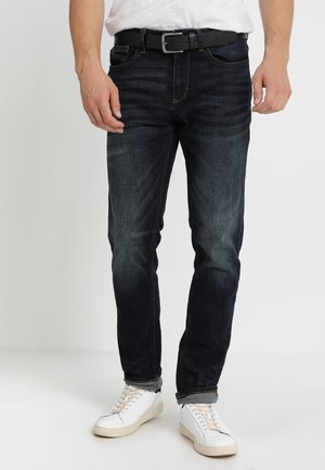 Homme portant un jean bleu foncé avec une ceinture noire, des baskets blanches et une chemise blanche rentrée, debout devant un fond uni.