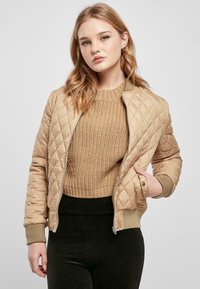 Beige quiltet bomberjakke med ribbede mansjetter og liv. Kombinert med en teksturert strikket beige genser og mørke tettsittende bukser.