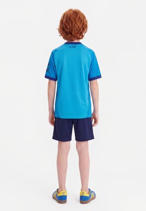 Enfant roux vu de dos portant un maillot de sport bleu, un short bleu marine et des baskets Adidas bleues et jaunes sur fond blanc.