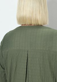 Grünes, strukturiertes Hemd mit gefalteter Rückendesign, Kragen und sichtbaren Nähdetails. Der Stoff hat ein zerknittertes Aussehen.