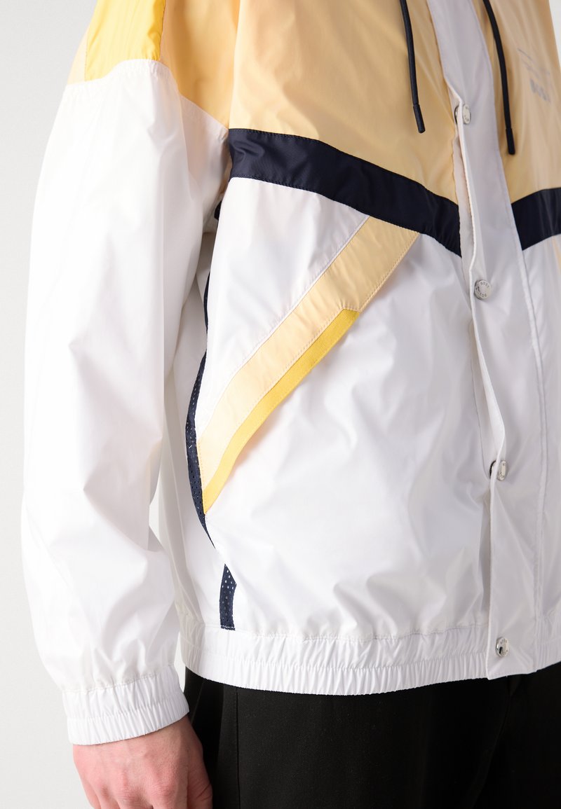 Veste coupe-vent colorée avec une base blanche et des accents jaunes et bleu marine, des poignets élastiques et un design texturé sur les manches.
