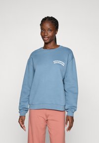 Kvinne med flette i håret iført en blå hettegenser med teksten "Peaceful Warrior" og løse rosa bukser, stående mot en ensfarget lys bakgrunn.