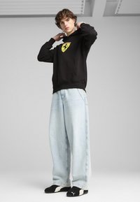 Sort hættebluse med gul Ferrari-logo, parret med lyseblå vide jeans og sorte sportssko. Casual, afslappet pasform.