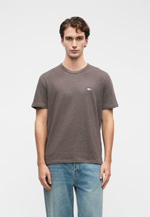 Junger Mann mit kurzen, lockigen Haaren, der ein braunes Kurzarm-T-Shirt und hellblaue Jeans trägt und vor einem weißen Hintergrund steht.