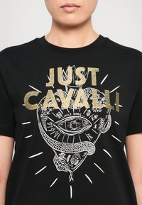 Black T-shirt with gold glitter "JUST CAVALLI" text over a white graphic of an eye and snake with small tattoo-style illustrations.
