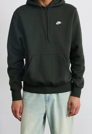 Persoon draagt een donker groene Nike hoodie met voorzak en lichtblauwe spijkerbroek, staand met ontspannen armen langs het lichaam.