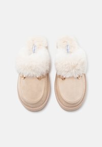 Beige slip-on hjemmesko med en blød, luksuriøs cremefarvet pelskant, udsmykket med guldfarvet hardware i overdelen. Struktureret ruskindsmateriale.