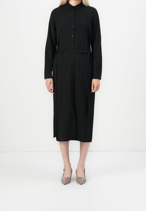 Kvinde iført en sort midi-kjole med lange ærmer, knapper, krave og matchende bælte, kombineret med taupe spidse hæle.