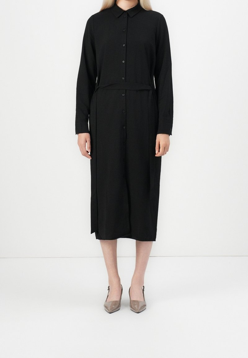 Femme portant une robe midi noire à manches longues, boutonnée, avec col et ceinture assortie, associée à des escarpins pointus taupe.