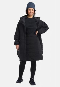 Giacca imbottita nera con cappuccio, maniche lunghe e tasche frontali, abbinata a una maglia nera, gonna e leggings. Berretto blu a coste indossato.