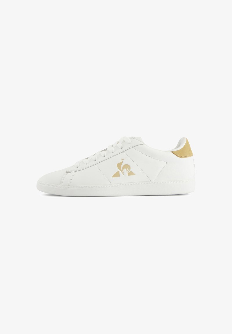 White leather sneaker with a textured surface, golden accent at the heel, and a logo on the side. Features flat laces and a rubber sole.