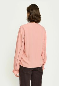 Rosa Bluse mit langen Ärmeln, gerafften Bündchen und dekorativen Nähten an den Seiten. Das Material wirkt glatt und leicht.