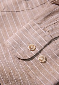 Camicia a righe marrone chiaro con linee verticali bianche. Tessuto morbido, con polsini bottonati e bottoni tondi di colore chiaro.