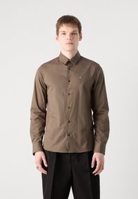 Camisa de manga longa castanha com colarinho abotoado, botões pretos e um pequeno logótipo no lado esquerdo do peito, combinada com calças escuras.