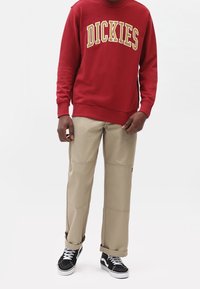 Felpa rossa con la scritta "DICKIES" in lettere gialle, abbinata a pantaloni beige con cuciture. Sneakers in bianco e nero completano l'outfit.