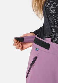 Persoon die de paarse tailleband van sneeuwbroek met klittenbandsluiting en ritssluiting aanpast, terwijl hij een zwart geblokt sweater met bretels draagt.