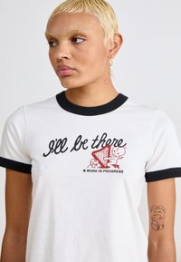 Witte katoenen T-shirt met zwarte ringerhals, voorzien van een grafische print van een personage dat harp speelt en de tekst "Ik ben er bij" in cursief.
