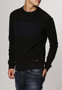 Sudadera negra con un logo texturizado "BOSS ORANGE" en azul oscuro, cuello redondo, puños y dobladillo de canalé. Combinada con jeans de mezclilla oscuros.