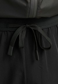 Schwarzer Kordelzug, der zu einer Schleife am Bund schwarzer Sporthosen gebunden ist, mit einem Teil einer Reißverschlussjacke darüber.