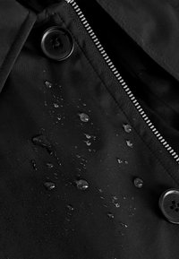 Gros plan d'un tissu noir avec des gouttes d'eau, des boutons noirs et une fermeture éclair argentée partiellement visible.