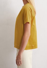 Haut moutarde à manches courtes avec une coupe ample, en tissu texturé. Encolure carrée et ourlet droit. Porté avec un pantalon de couleur claire.