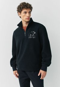 Schwarzer Fleece-Pullover mit einem Viertelreißverschluss-Kragen, strukturiertem Stoff, gesticktem Cartoon-Design auf der linken Brust und passgenauen Bündchen.