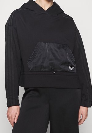 Czarna bluza z kapturem z błyszczącą przednią kieszenią na suwak i subtelnymi paskami na rękawach, noszona z czarnymi spodniami, bez widocznej twarzy ani szczegółów tła.