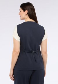 Navyblauwe top met korte crème mouwen, voorzien van een strikdetail aan de achterkant met een zilveren ring en een gladde, zachte textuur.