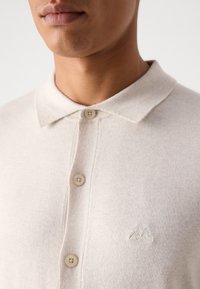 Beige kokvilnas polo krekls ar apkakles kakla izgriezumu, trim pogām, teksturētu audumu un mazu izšūtu logotipu uz krūšu daļas.