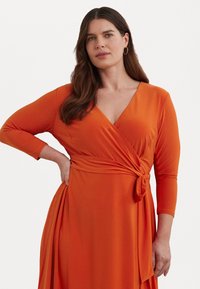 Oranža apvalka kleita ar garām piedurknēm un V veida kakla izgriezumu. Izceļas ar jostas siksnu un plūstošu svārku ar sānu izgriezumu. Gluda auduma tekstūra.