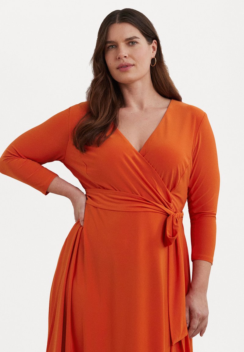 Oranža apvalka kleita ar garām piedurknēm un V veida kakla izgriezumu. Izceļas ar jostas siksnu un plūstošu svārku ar sānu izgriezumu. Gluda auduma tekstūra.