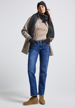 Femme portant un manteau beige, un pull beige, un jean bleu, des gants noirs, une écharpe, un chapeau et des bottes en daim beige, se tenant devant un fond gris clair.