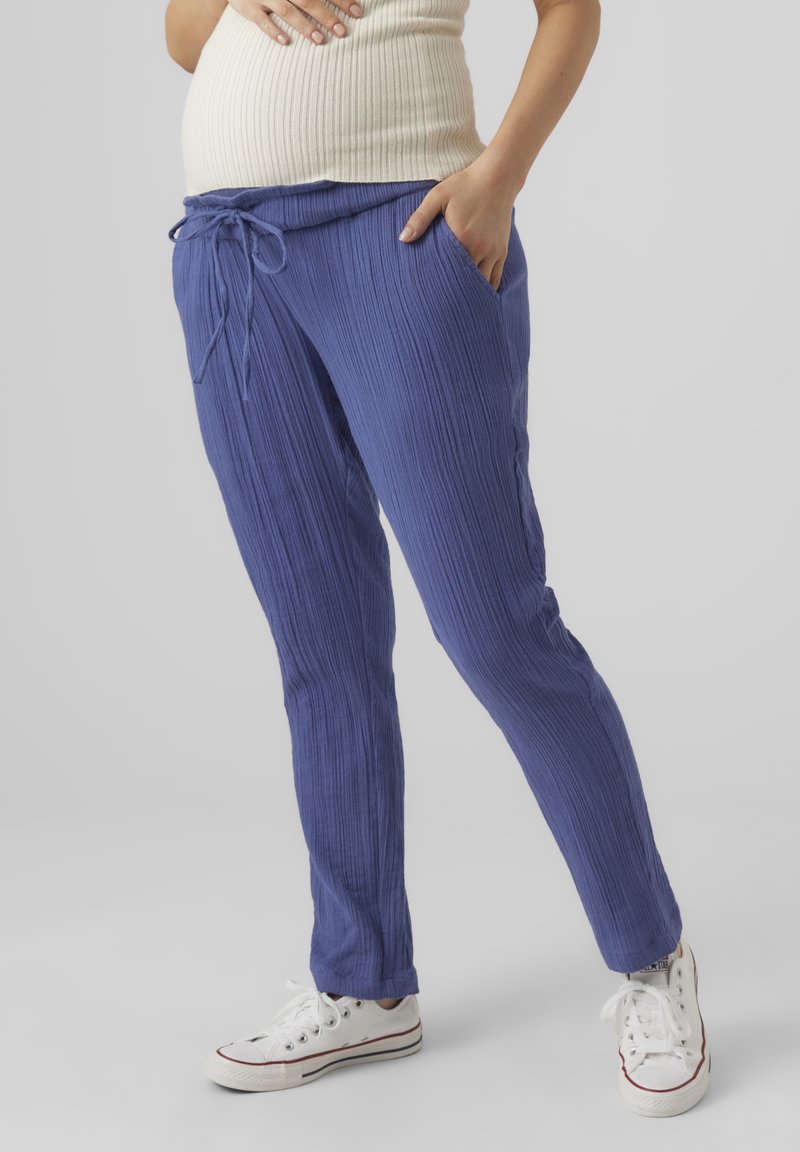 Une personne enceinte portant un pantalon de maternité violet côtelé avec cordon de serrage et des baskets blanches, une main reposant sur le ventre et l'autre dans la poche.