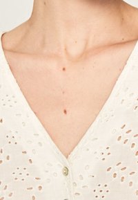 Bílý lněný top s výstřihem do V a výšivkou dírek, zdobený malými kulatými knoflíky a jemnými texturovanými vzory.