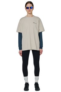 Vrouw draagt beige oversized T-shirt over een donkerblauw top met lange mouwen, zwarte leggings, witte sokken, zwarte sneakers en reflecterende sportzonnebril.