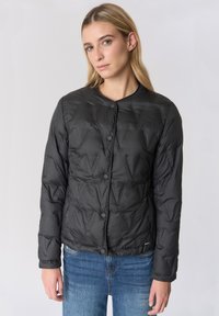 Giacca imbottita nera trapuntata con scollo rotondo, chiusura a bottoni a pressione e superficie testurizzata, abbinata a jeans blu.