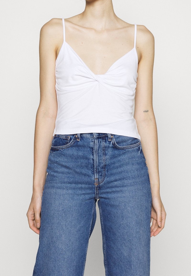 Valkoinen napakka toppi, jossa on syvä V-pääntie ja kierretyt yksityiskohdat edessä, yhdistettynä korkeavyötäröisiin sinisiin farkkuihin. Spagettihihnat ovat näkyvissä.