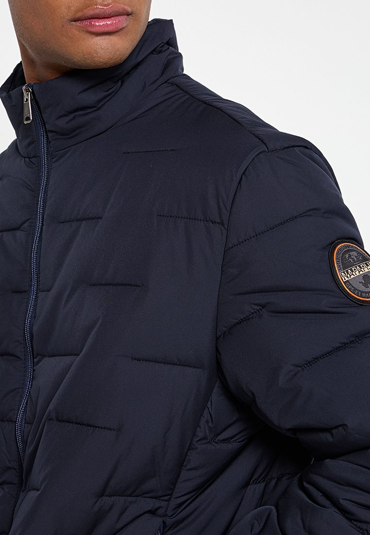 Dunkelblaues Daunenjacke mit hohem Kragen, gequilteten Linien, einem Frontreißverschluss und einem runden Logo-Patch auf der linken Schulter.