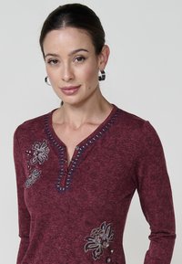 Burgundijas garām piedurknēm krekls ar izgrieztiem apkakli, ar ziliem akcentiem, ziedu izšuvumu un pogu detaļām priekšpusē.