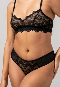 Svart spetstopp med vågiga kanter och tunna axelband, tillsammans med matchande genomskinliga spetstrosor med ett blommönster. Slät textur.