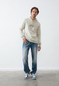 Light beige hoodie with logo, cotton blend, drawstring hood. Worn with faded blue jeans, sneakers with teal and white accents.