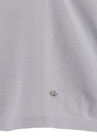 Camiseta de algodón gris claro con una textura suave, que presenta una pequeña etiqueta de logo plateada en el dobladillo. Diseño simple, de manga corta.