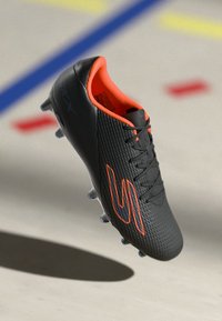 Svart fotballstøvel med teksturert overflate, oransje detaljer og fremtredende logo. Har tradisjonelt snøring og formsydde knotter for godt grep.