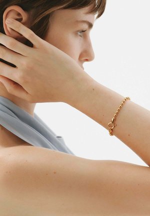 Pulsera de cuentas de oro con un cierre simple, llevada en la muñeca de un modelo, con piel suave y una bufanda azul claro de fondo.