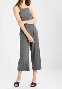 Mono gris sin mangas con un cuerpo ajustado y piernas anchas y cortas. Hecho de una tela suave con una textura lisa. Sandalias planas negras.